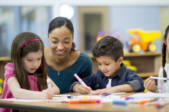 A multi-ethnic group of elementary age children are coloring together at their desk. Their female teacher is sitting next to them and giving them encouragement.