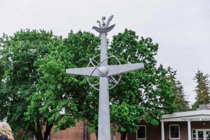 A cross statue with the lamp of learning lit at the top of the cross. With trees and a building in the abckground.