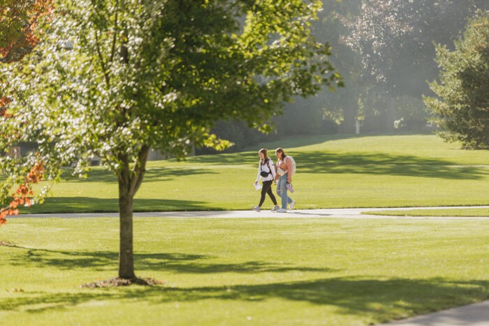 Students walking across campus with dew on the grass. There are trees and fresh, green grass with the sun coming up.